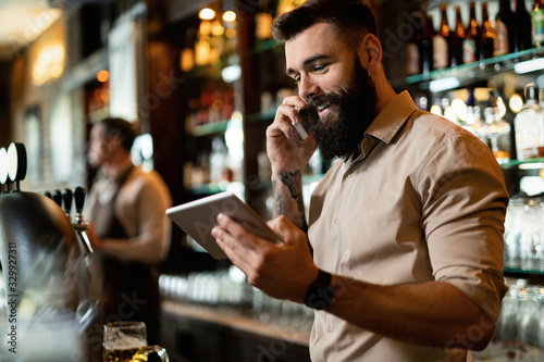 Young happy bartender using touchpad while talking on the phone.