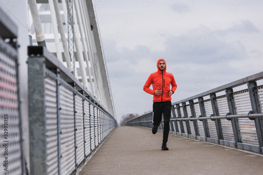 Handsome young athlete running fast along big modern bridge in orange windbreaker jacket. Exercising, Jogging, Sport, Winter. Male athlete running.