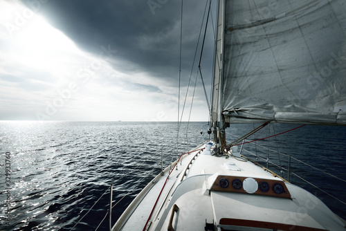 White sloop rigged yacht sailing near the coast of Maine, Southwest Harbor, USA. A view from the deck to the bow, mast and sails. Dark blue storm sky, sun rays through the clouds