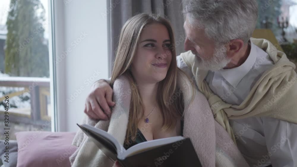 Senior Caucasian man in love talking with young pretty Caucasian woman reading book. Happy loving couple with age difference enjoying time together indoors. Lifestyle, love, romance, tranquility. Senior Caucasian man in love talking with young pretty Caucasian woman reading book. Happy loving couple with age difference enjoying time together indoors. Lifestyle, love, romance, tranquility.