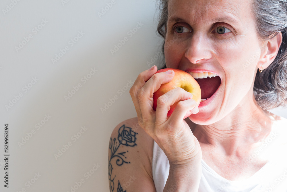 Close-up Of Mature Woman Eating Apple Against White Wall - obrazy, fototapety, plakaty 