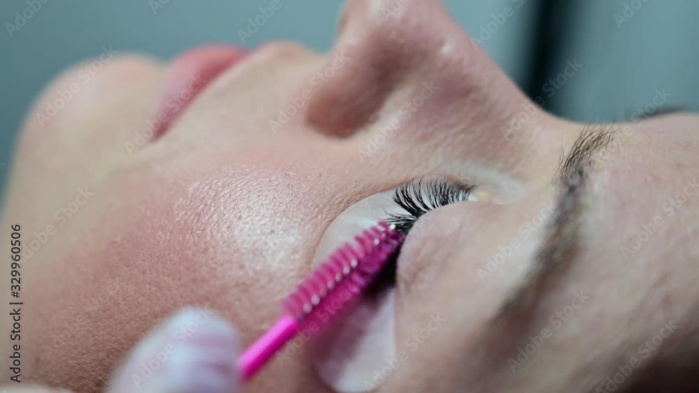 Close-up profile of a woman lying with her eyes closed. The master combs the extension eyelashes with a pink brush. Permanent makeup. Close-up profile of a woman lying with her eyes closed. The master combs the extension eyelashes with a pink brush. Permanent makeup.