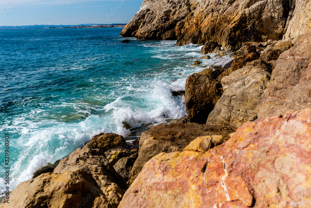The Mediterranean Sea waves reaching the big rocks on the shore with the sunlight reflecting in the turquoise water on a sunny day (Nice, France)