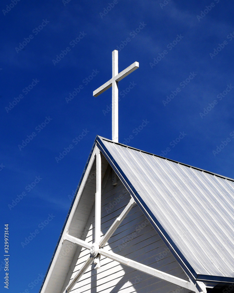 A simple cross stretching into a blue sky from the top of a small town church of white washed boards and tin roof. A simple cross stretching into a blue sky from the top of a small town church of white washed boards and tin roof.