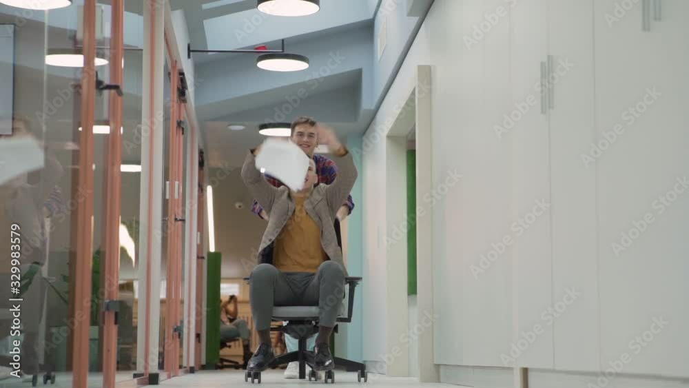 Ecstatic young man riding chair down office corridor being pushed by male colleague, shouting happily and tossing papers in air in slow motion celebrating achievement Ecstatic young man riding chair down office corridor being pushed by male colleague, shouting happily and tossing papers in air in slow motion celebrating achievement