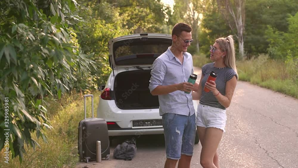 Portrait of a couple in love with thermo mugs near the car on the road in the woods, near the open trunk is a suitcase and a bag. Summer trip by car. Portrait of a couple in love with thermo mugs near the car on the road in the woods, near the open trunk is a suitcase and a bag. Summer trip by car.