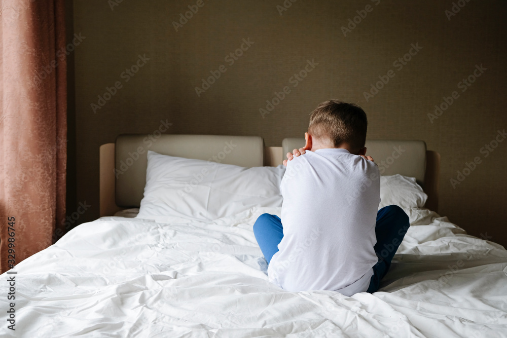 Unhappy offended little boy sitting alone on the bed, back to camera. Loneliness, variance, autism concept Unhappy offended little boy sitting alone on the bed, back to camera. Loneliness, variance, autism concept