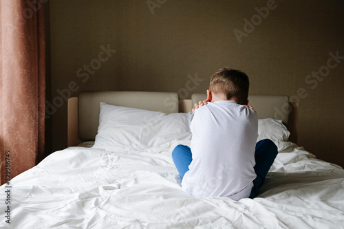 Unhappy offended little boy sitting alone on the bed, back to camera. Loneliness, variance, autism concept