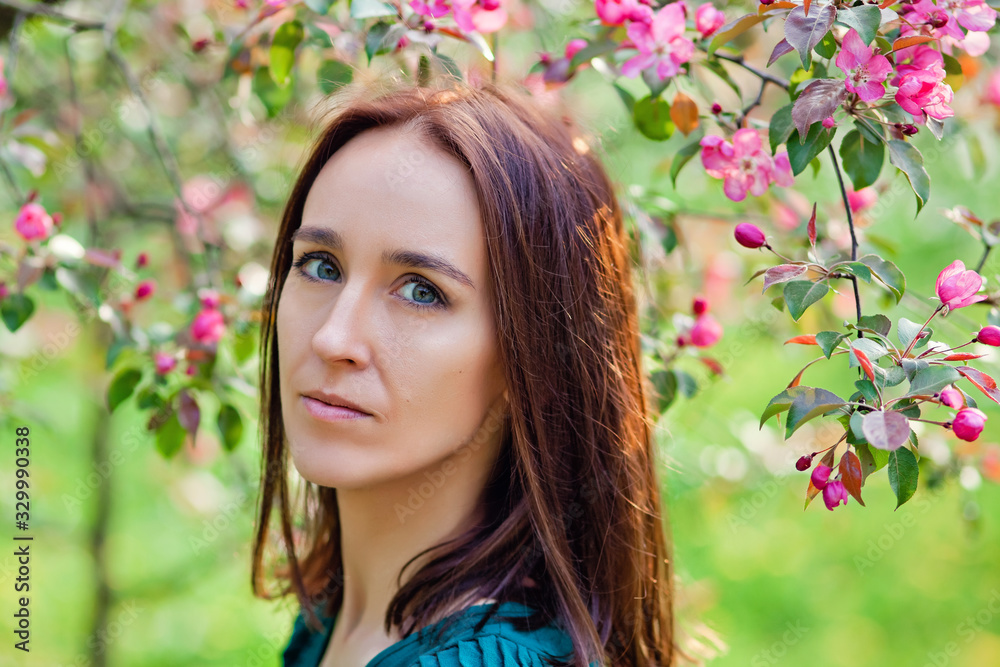 Fototapeta premium A beautiful young brunette stands near a blooming Apple tree on a warm spring day. Springtime