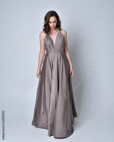  Portrait of a pretty brunette girl wearing a long silver evening gown, full length standing pose against a studio background.