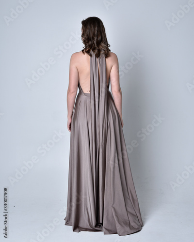  Portrait of a pretty brunette girl wearing a long silver evening gown, full length standing pose against a studio background.