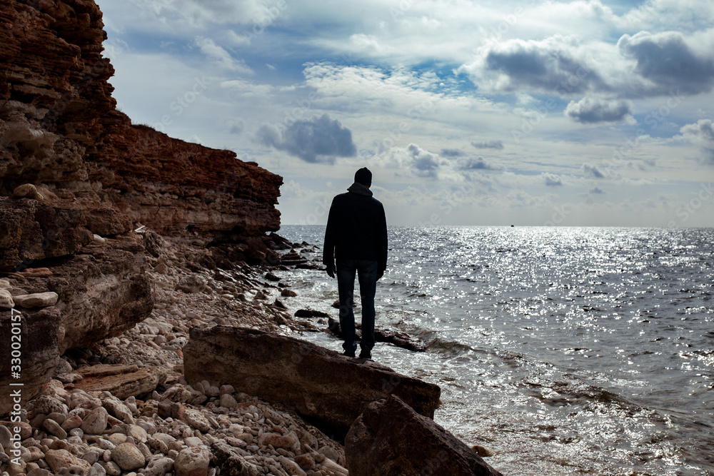 adult man looks at the sea, he stands with his back and is dressed in a jacket and jeans, a walk along the sea shore in spring, silhouette