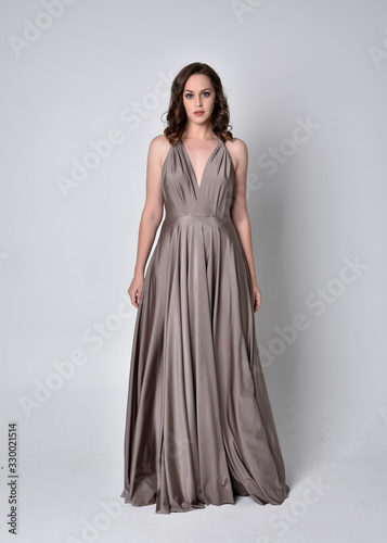  Portrait of a pretty brunette girl wearing a long silver evening gown, full length standing pose against a studio background.
