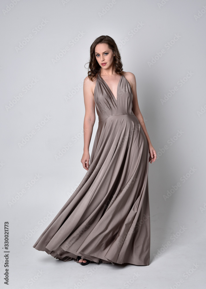  Portrait of a pretty brunette girl wearing a long silver evening gown, full length standing pose against a studio background.