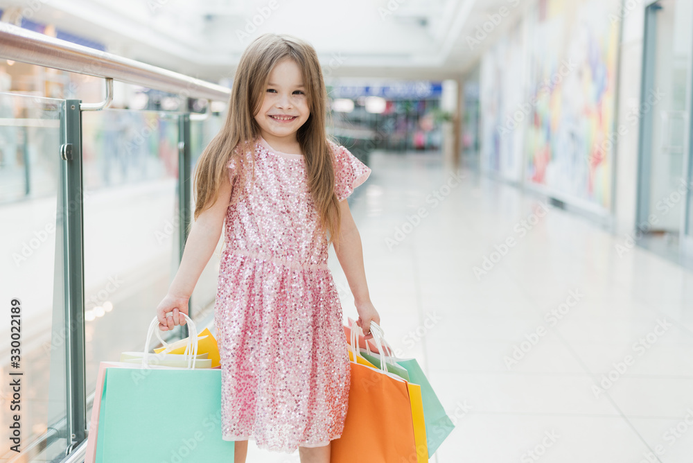 Portrait of a little happy girl in the mall. A smiling laughing girl in a pink dress with multi-colored bags in her hands is engaged in shopping. Template for your advertisement Portrait of a little happy girl in the mall. A smiling laughing girl in a pink dress with multi-colored bags in her hands is engaged in shopping. Template for your advertisement
