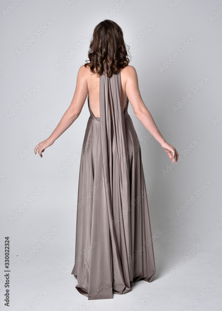  Portrait of a pretty brunette girl wearing a long silver evening gown, full length standing pose against a studio background.