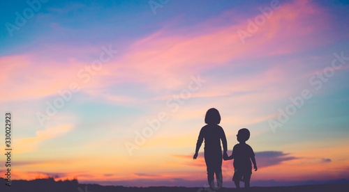 Tableau sur toile Silhouette back view of two children sibling walking on the beach while holding hands with sunset background