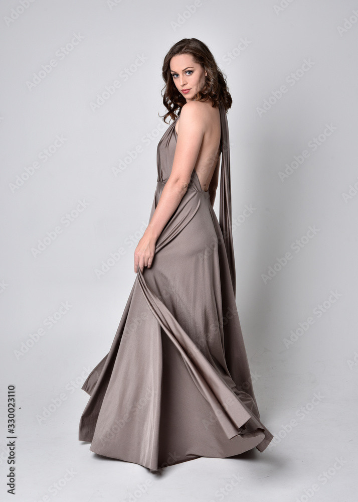 Portrait of a pretty brunette girl wearing a long silver evening gown, full length standing pose against a studio background. Portrait of a pretty brunette girl wearing a long silver evening gown, full length standing pose against a studio background.