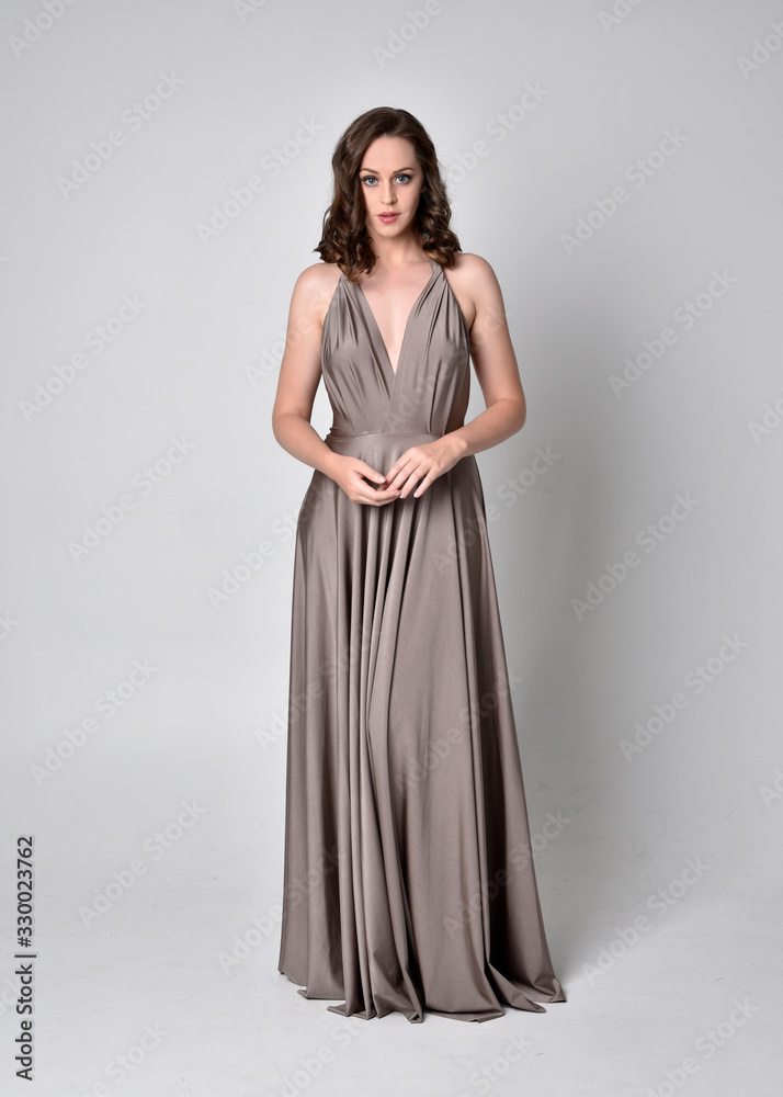 Portrait of a pretty brunette girl wearing a long silver evening gown, full length standing pose against a studio background. Portrait of a pretty brunette girl wearing a long silver evening gown, full length standing pose against a studio background.