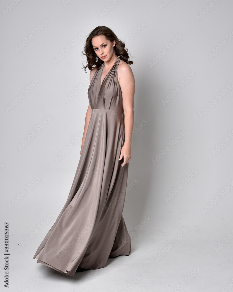  Portrait of a pretty brunette girl wearing a long silver evening gown, full length standing pose against a studio background.