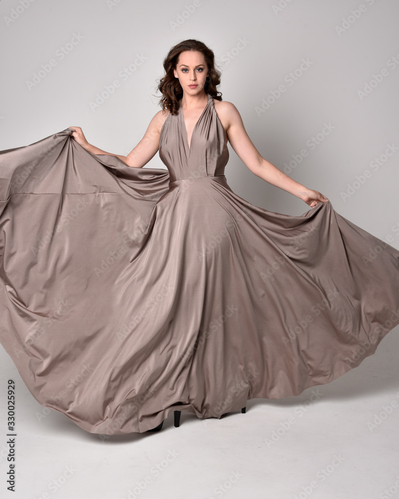 Portrait of a pretty brunette girl wearing a long silver evening gown, full length standing pose against a studio background. Portrait of a pretty brunette girl wearing a long silver evening gown, full length standing pose against a studio background.