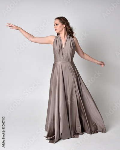  Portrait of a pretty brunette girl wearing a long silver evening gown, full length standing pose against a studio background.