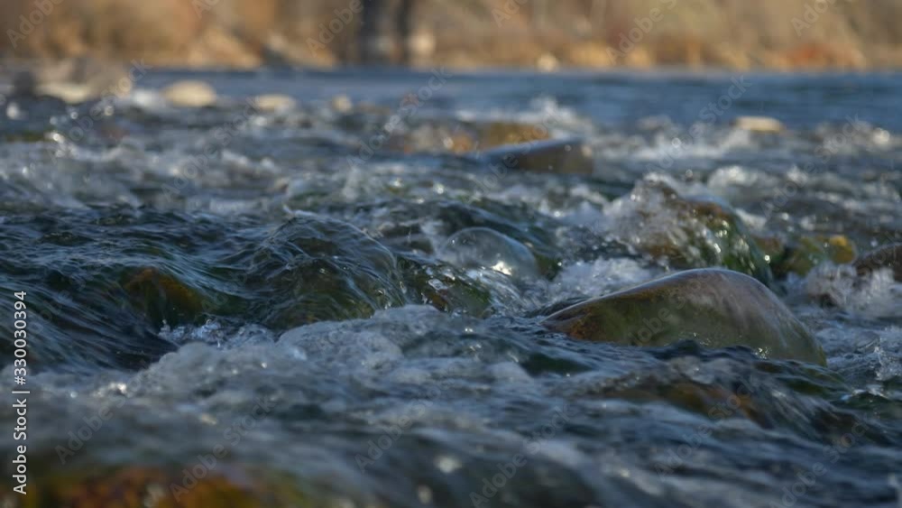 Low angle slow motion slider pulling back over small mossy rock river rapids at sunset/golden hour. River bank in golden hour light in background. Low angle slow motion slider pulling back over small mossy rock river rapids at sunset/golden hour. River bank in golden hour light in background.