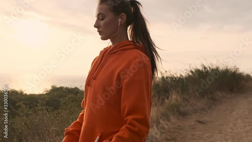 Concentrated pretty athletic woman in earphones warming up with legs and looking away at seashore photo