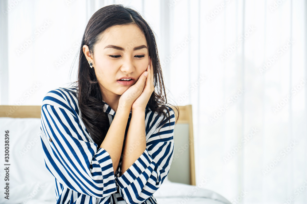 Young Asian woman touching cheek while suffering from toothache and discomfort on bed in white bedroom morning.Concept of women's health care. Young Asian woman touching cheek while suffering from toothache and discomfort on bed in white bedroom morning.Concept of women's health care.