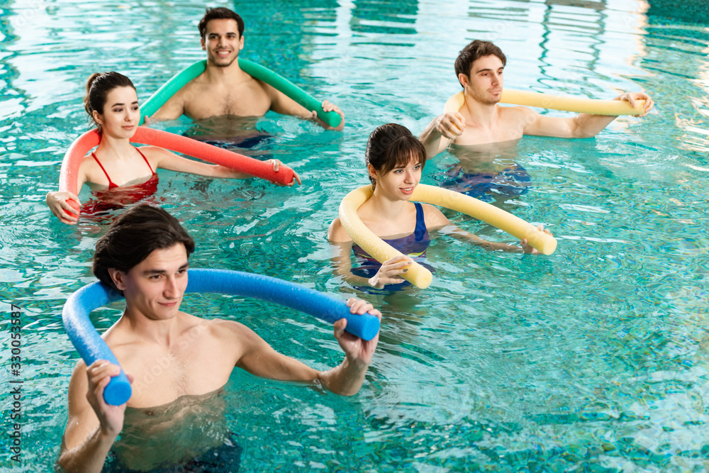 Smiling young people exercising with pool noodles during water aerobics in swimming pool