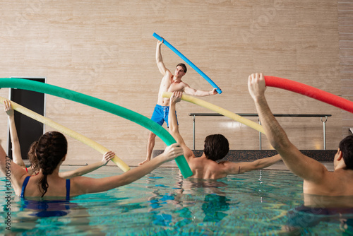 Fototapeta Naklejka Na Ścianę i Meble -  Selective focus of group of people holding pool noodles while exercising with trainer in swimming pool