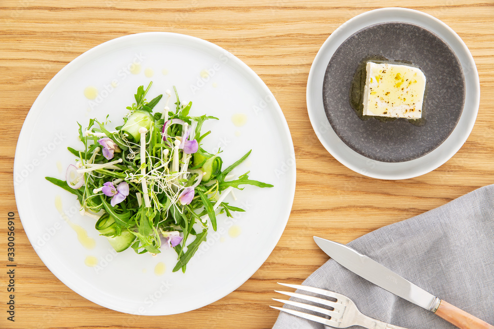 Arugula salad with bean sprout, enoki mushrooms, cucumber and edible flowers, feta cheese on the side over a wood background. Napkin, knife and fork. Flat lay. Vegetarian recipe. Arugula salad with bean sprout, enoki mushrooms, cucumber and edible flowers, feta cheese on the side over a wood background. Napkin, knife and fork. Flat lay. Vegetarian recipe.