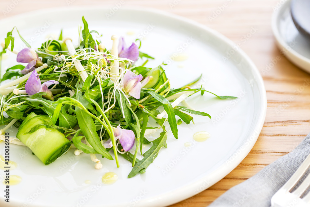 Arugula salad with bean sprout, enoki mushrooms, cucumber and purple edible flowers, over an oak wood background. Grey napkin, and silver fork. Vegetarian recipe. Arugula salad with bean sprout, enoki mushrooms, cucumber and purple edible flowers, over an oak wood background. Grey napkin, and silver fork. Vegetarian recipe.