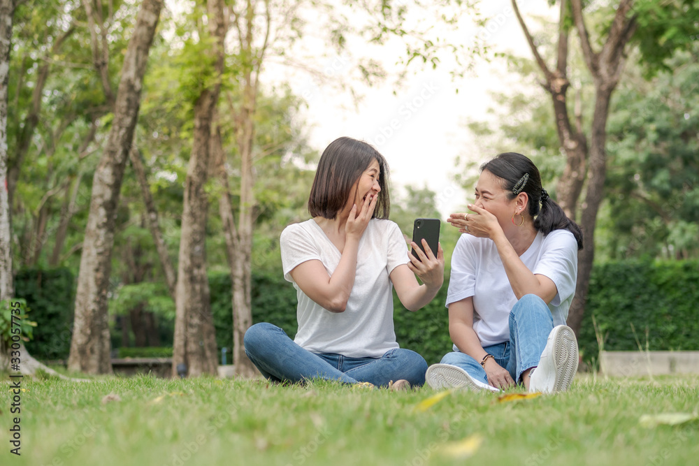 Two happy young girl friends sitting on grass in a park. Looking at smartphone.
