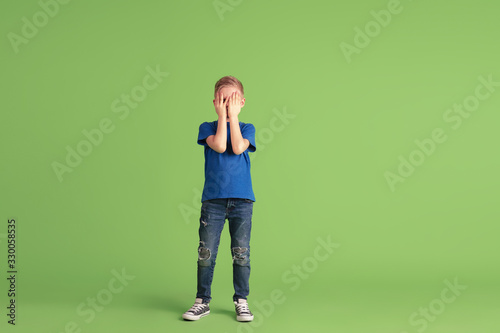 Covering face, looking throught hand. Happy boy playing and having fun on green studio background. Caucasian kid looks playful, smiling. Concept of education, childhood, emotions, facial expression. © master1305