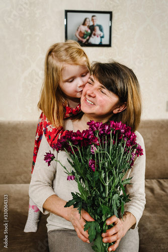Little girl making surprise for grandmother, congratulating her with birthday. Happy grandmother and granddaughter embracing and kiss together at home. Women's day. Grandma holding bouquet flowers.