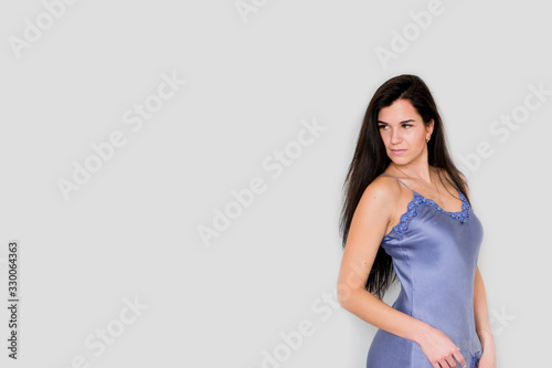 Portrait sexy beautiful serious young woman with long straight dark hair in blue silk nightdress with lace trim on grey background wall. Caucasian Female looking away. Copy space for text.
