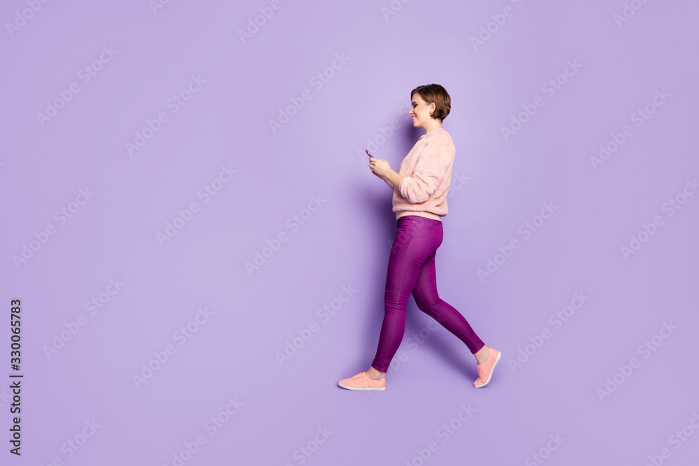 Full length profile photo of pretty lady holding telephone chatting with friends walking down street wear casual stylish pink pullover trousers isolated purple color background