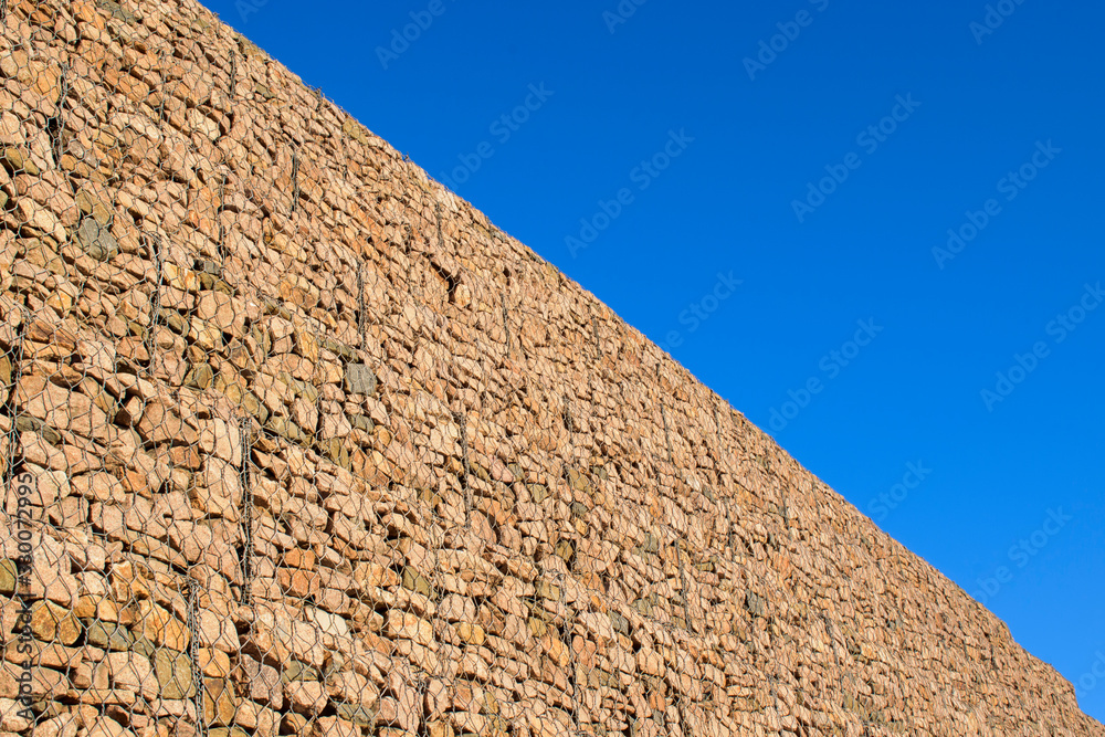 High wall of gabions. Stones in a metal mesh. Blue sky background.