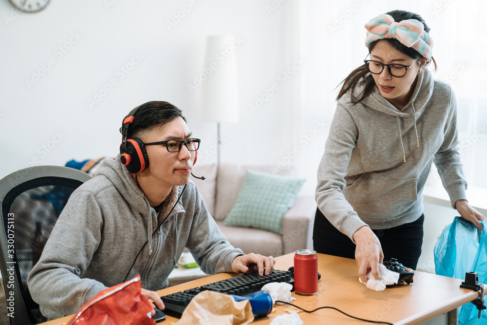 Lazy asian guy playing online video game on computer while his girlfriend  cleaning table at home. frowning woman murmur at her boyfriend. man in  headphones concentrated having fun relax on technology Stock