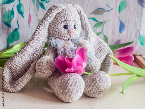 Knitted light gray handmade crafted rabbit with pink tulips .