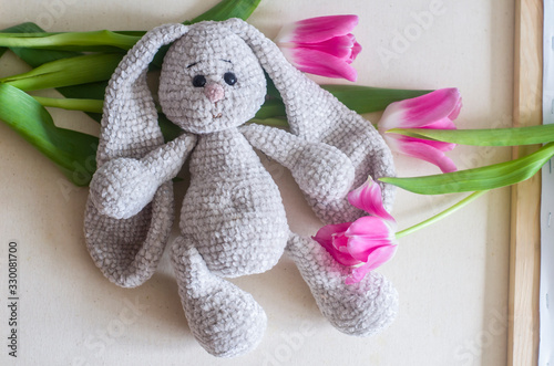 Knitted light gray handmade crafted rabbit with pink tulips .