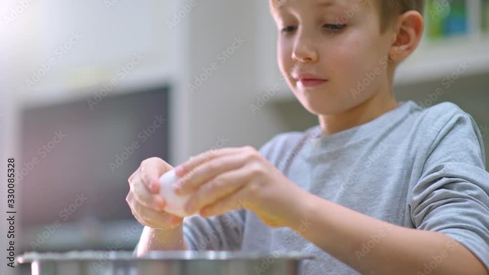 White boy takes an egg and break it to the bowl  with dough and then mixing.  Child cooking at the kitchen. Kid stirring ingredients for a cake in the steel bowl. Real time 4k footage