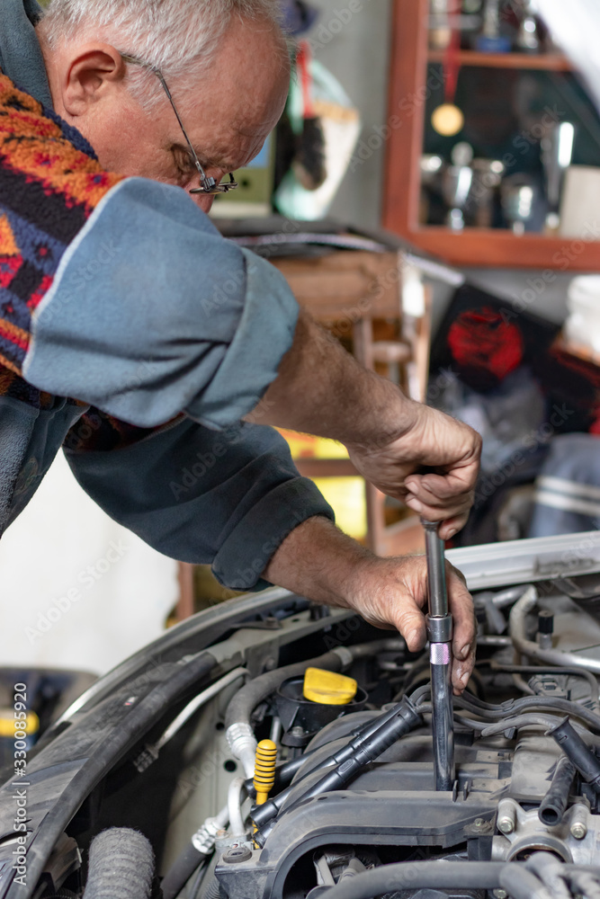 Portrait of an old car mechanic changing spark plugs on his old car in the garage. Fixing spark plugs with special long screwdriver. Old car mechanics and repair concept. Manual work. Portrait of an old car mechanic changing spark plugs on his old car in the garage. Fixing spark plugs with special long screwdriver. Old car mechanics and repair concept. Manual work.