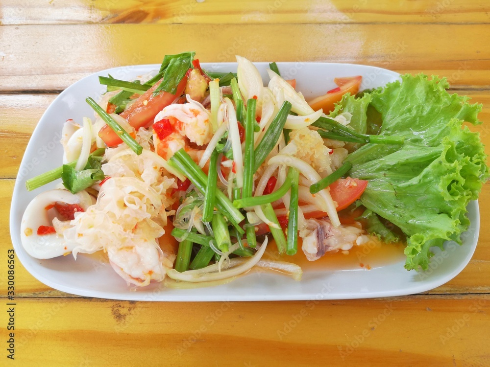 Thai food, mixed seafood salad Consists of squid, shrimp in a white plate With green vegetables beside There is a wooden floor in the background. Thai food, mixed seafood salad Consists of squid, shrimp in a white plate With green vegetables beside There is a wooden floor in the background.