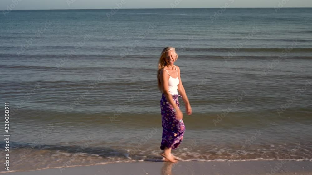 Playful mature woman wearing white top and batik skirt walking along the shore at a resort with arms raised and smiling at camera. Playful mature woman wearing white top and batik skirt walking along the shore at a resort with arms raised and smiling at camera.