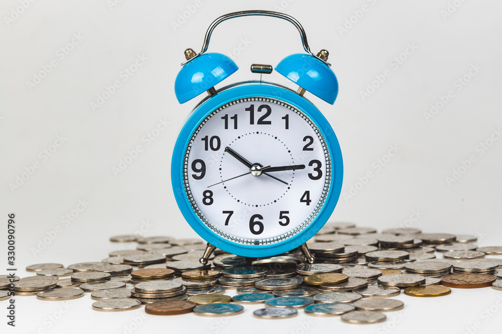 Blue analog clock alarm over money coins against white background - Time is Money concept Blue analog clock alarm over money coins against white background - Time is Money concept