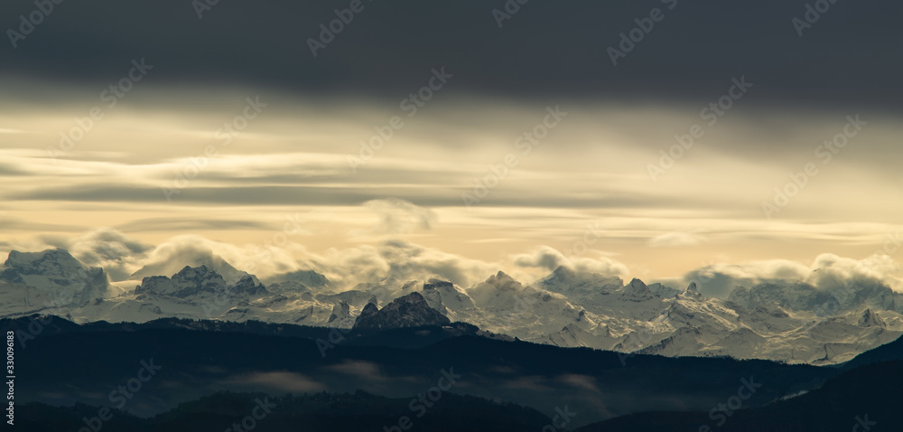 Dramatic landscape of snowy mountain tops in the Swiss alps as seen from the Zurich area late afternoon sun light illuminating from behind a thick sheet of clouds