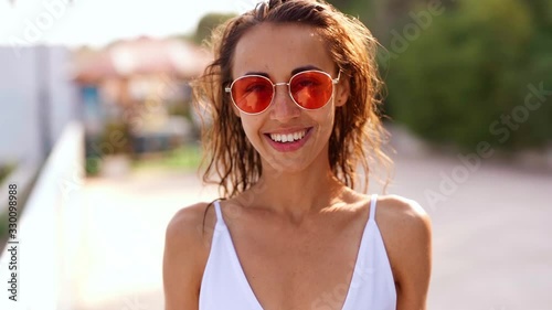 joyfully smiling Beautiful young tanned multyethnic woman in white bikini and pink sunglasses standing on bay at seaside on resort and smiling. slow motion footage photo