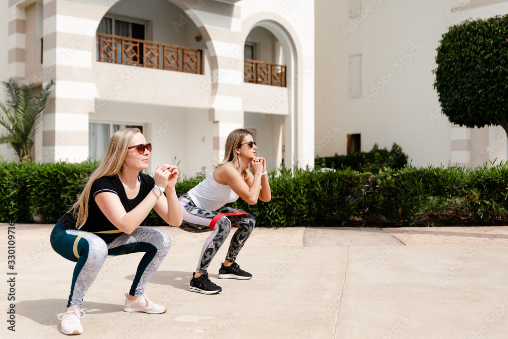 Obraz premium two young sports girls doing exercises with elastic,sportswoman portrait,stretch elastic squats